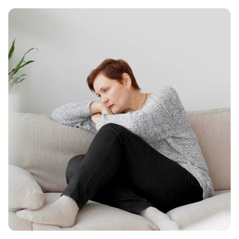 Older woman sitting on a couch hugging her knees, depicting fatigue, mood changes, or hormonal imbalance distress.